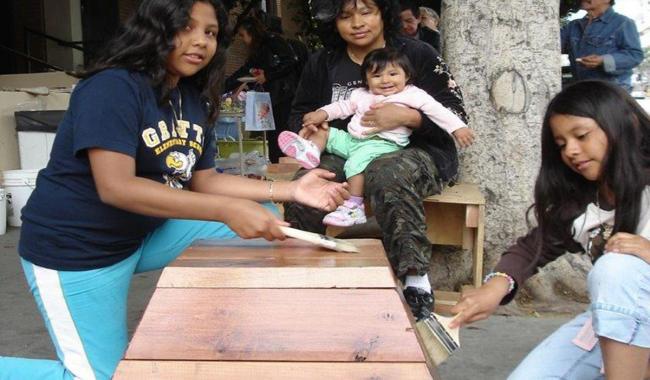 A mother sitting on a bench holding an infant in her arms and two girls painting a wooden bench with paint brushes